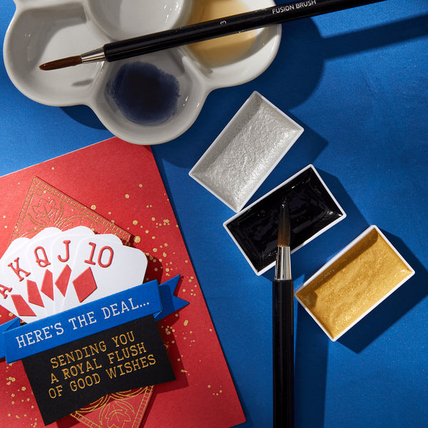 A water color mixing set with three watercolor paints in gold, silver, and indigo colors, displayed alongside a watercolor brush and a card with playing card symbols.